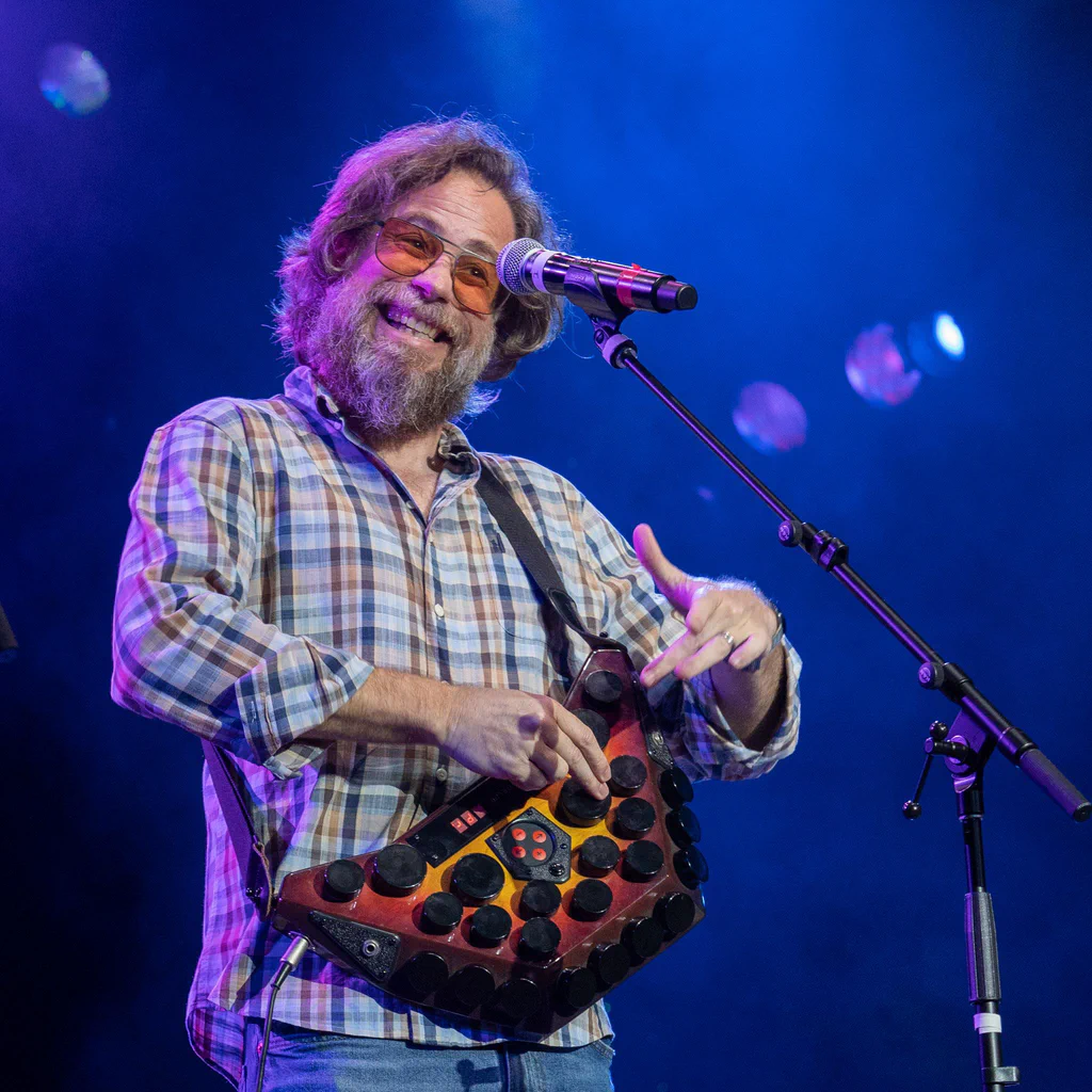 A man with glasses and a beard, smiling on stage with a colorful instrument, in front of a microphone at JoCo Cruise 2025.