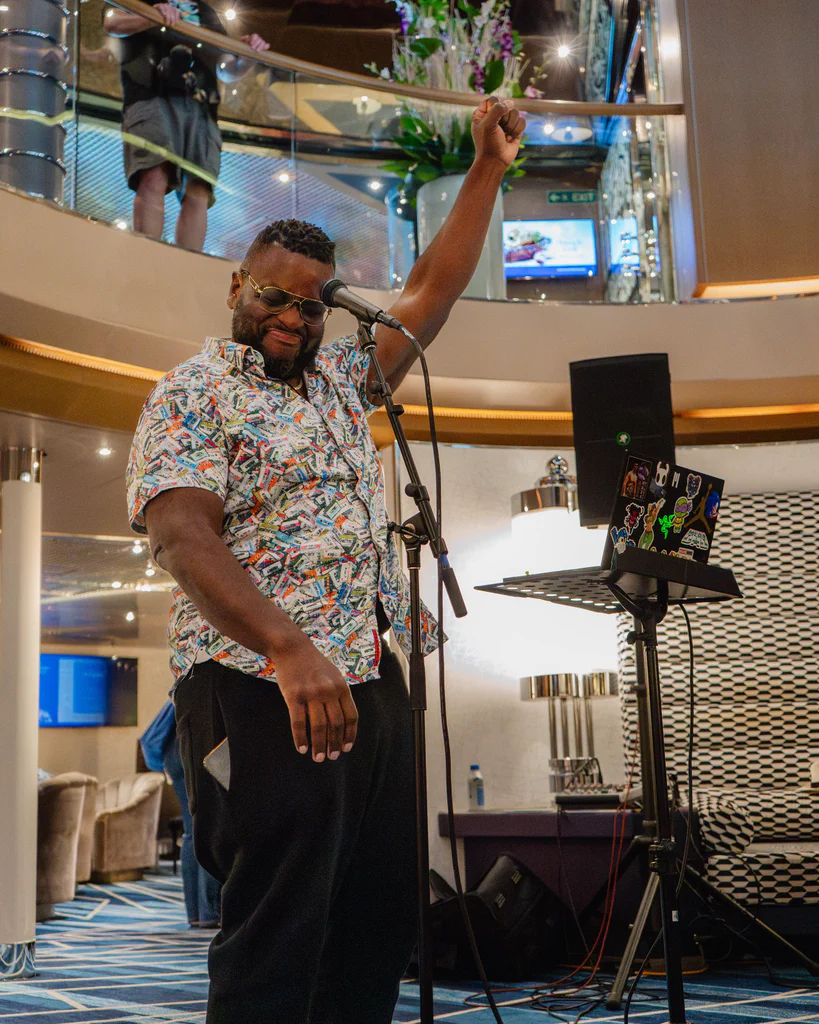 A performer with glasses and a colorful shirt raises one arm while singing into a microphone on JoCo Cruise 2025.