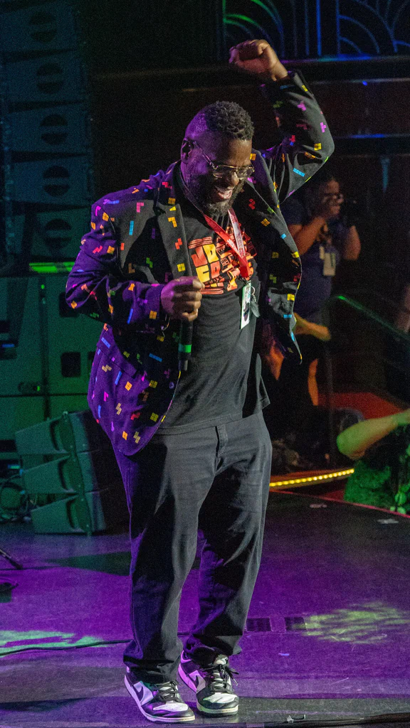 A man is dancing and smiling on stage during JoCo Cruise 2025, wearing a colorful, patterned blazer and sneakers.
