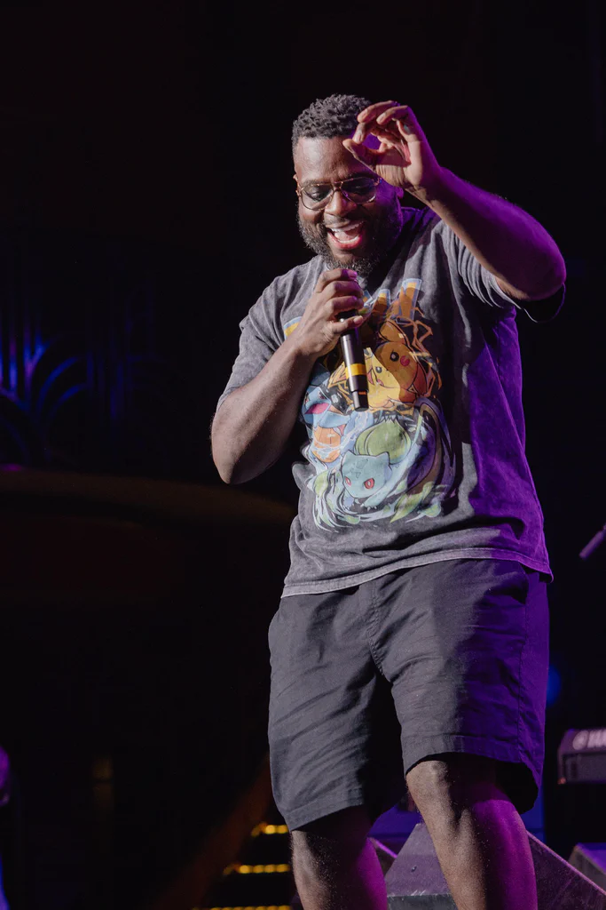A man singing on stage during JoCo Cruise 2025, holding a microphone and wearing a Pokémon-themed T-shirt.
