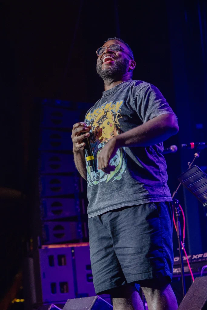 A man on stage smiling and holding a microphone, wearing a Pokémon T-shirt, with audio equipment in the background.