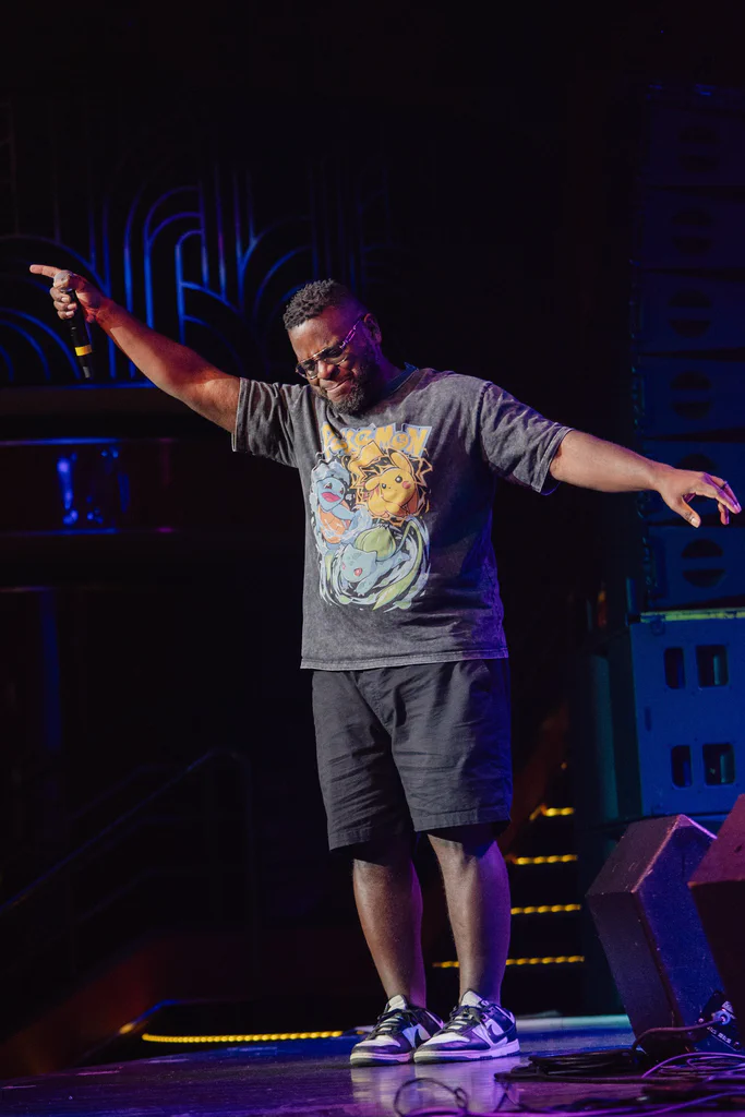 A man on stage at JoCo Cruise 2025, holding a microphone with his arms outstretched and smiling.
