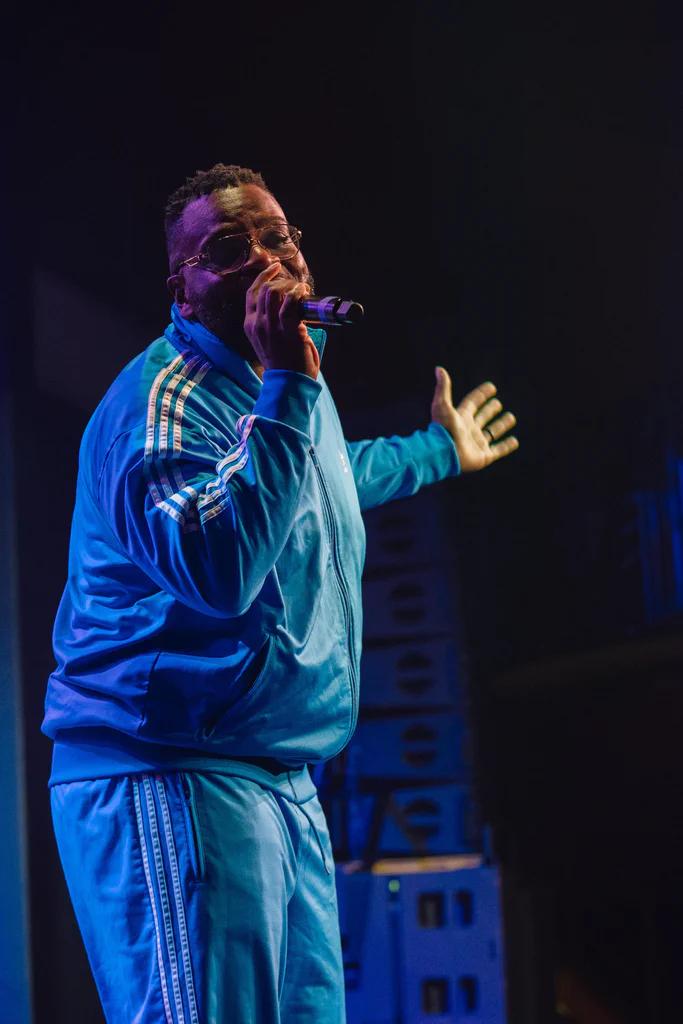 A man dressed in a blue tracksuit performs on stage at JoCo Cruise 2025, holding a microphone and gesturing with one hand.