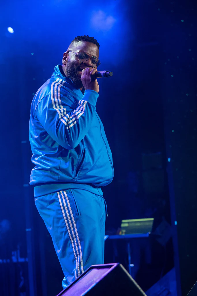A performer wearing a blue tracksuit with white stripes sings into a microphone on stage at JoCo Cruise 2025.