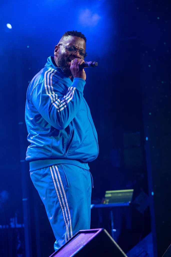 A performer wearing a blue tracksuit with white stripes sings into a microphone on stage at JoCo Cruise 2025.