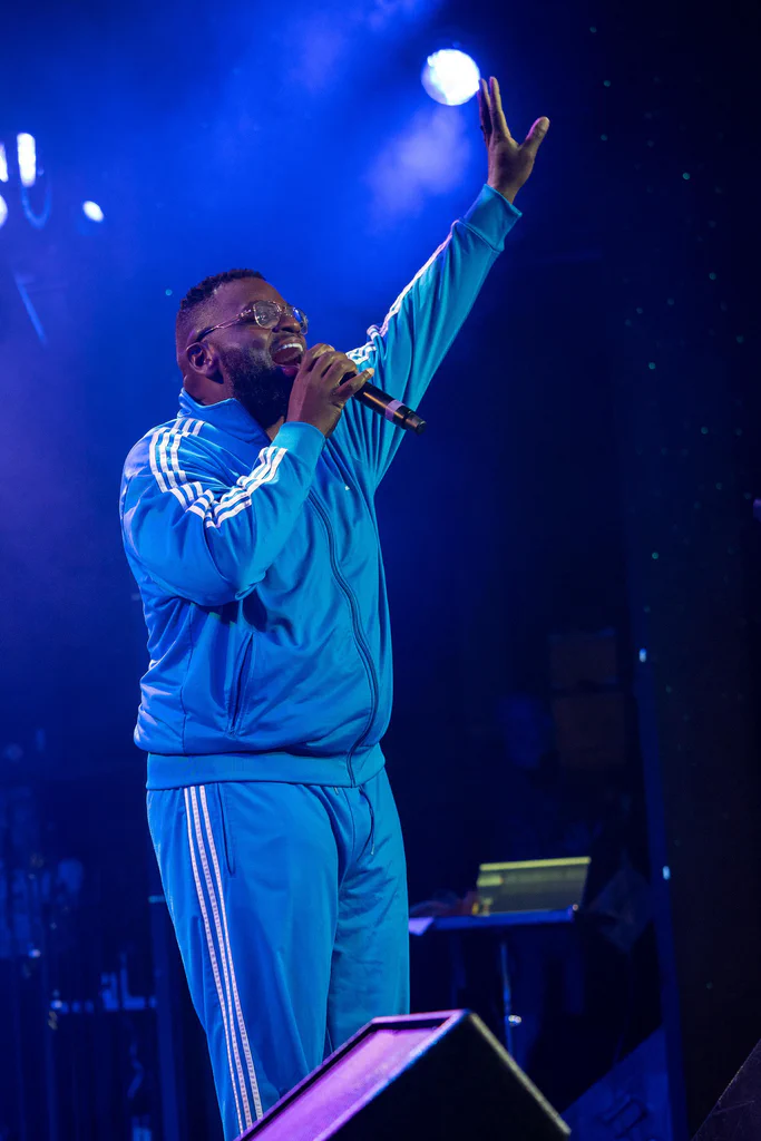 A performer in a blue tracksuit singing into a microphone on stage during JoCo Cruise 2025.