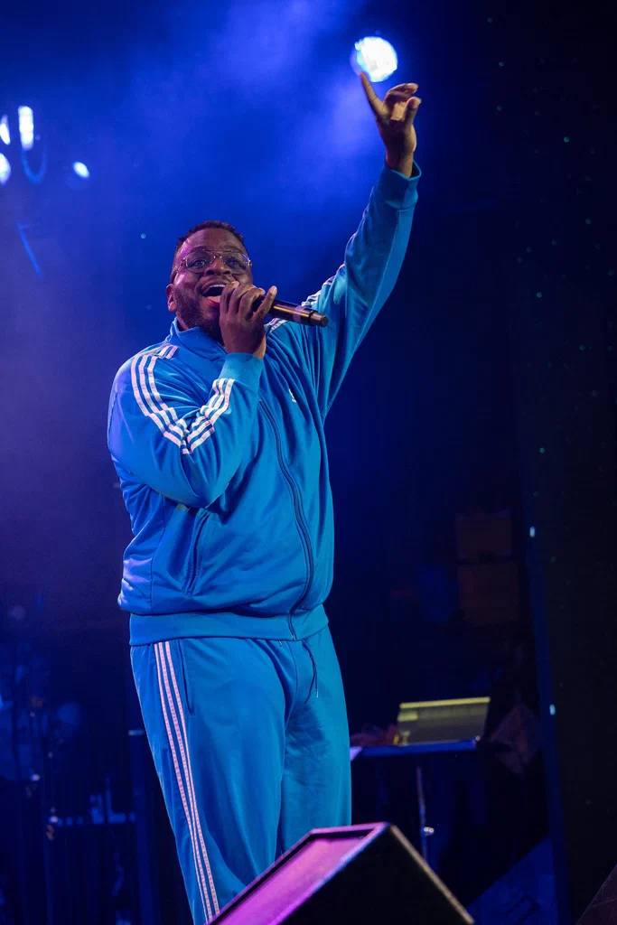A performer dressed in a blue tracksuit singing into a microphone with one arm raised on stage at JoCo Cruise 2025.