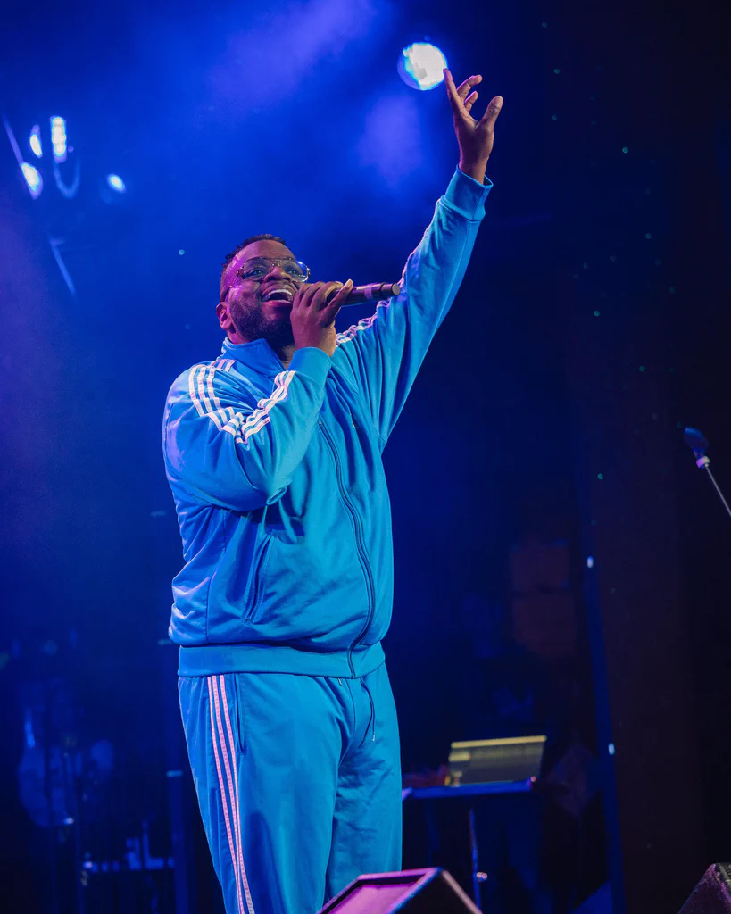 A man in a light blue tracksuit singing or speaking into a microphone on stage, with his hand raised and a spotlight illuminating him.