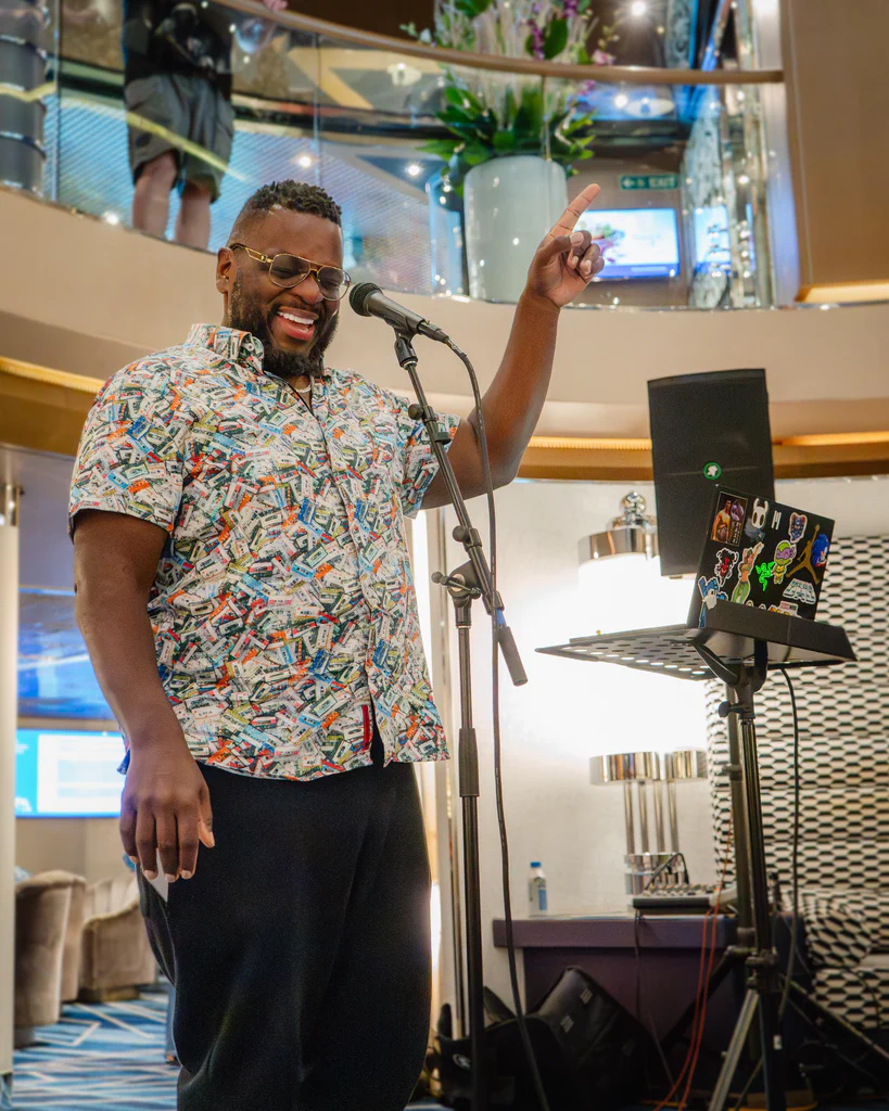 A man wearing sunglasses and a colorful patterned shirt is speaking or performing into a microphone on JoCo Cruise 2025, smiling and raising one hand.