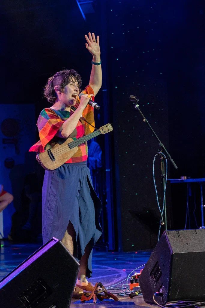 A woman on stage wearing colorful clothing, holding a ukulele, singing into a microphone, with her left arm raised in the air.