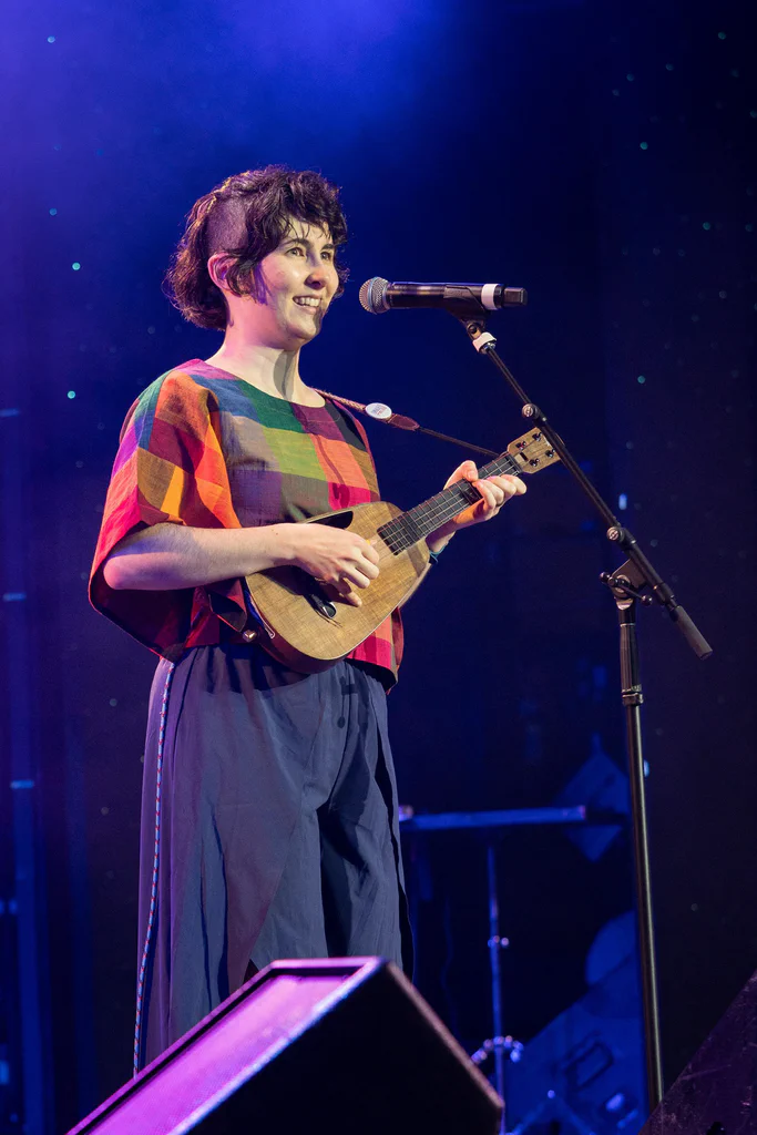 A woman with short curly hair plays a small ukulele and sings into a microphone on stage at JoCo Cruise 2025.