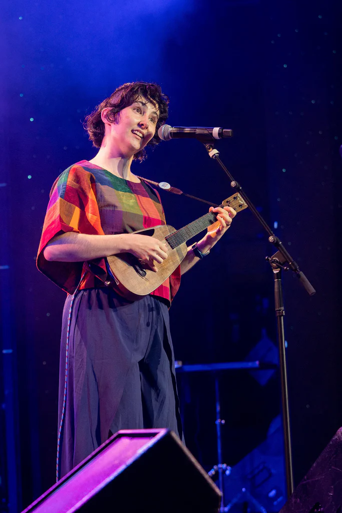 A woman performing on stage with a ukulele, singing into a microphone, wearing a colorful plaid top, with a dark starry background.