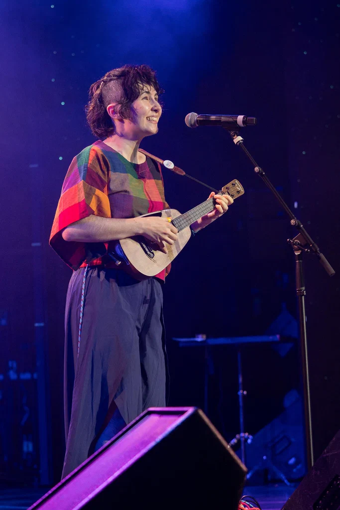 A woman with short, curly hair is performing on stage with a ukulele, smiling and singing into a microphone during JoCo Cruise 2025.