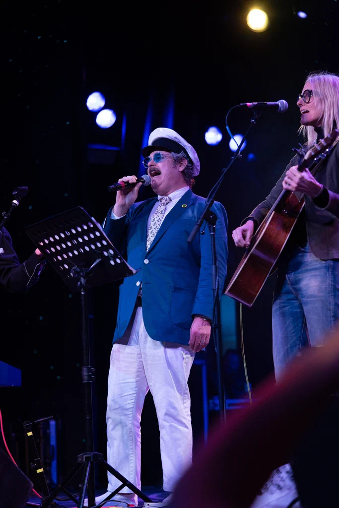 A man in a blue blazer, white pants, and a white sailor hat sings into a microphone while a woman next to him plays an acoustic guitar on stage during a performance at JoCo Cruise 2025.