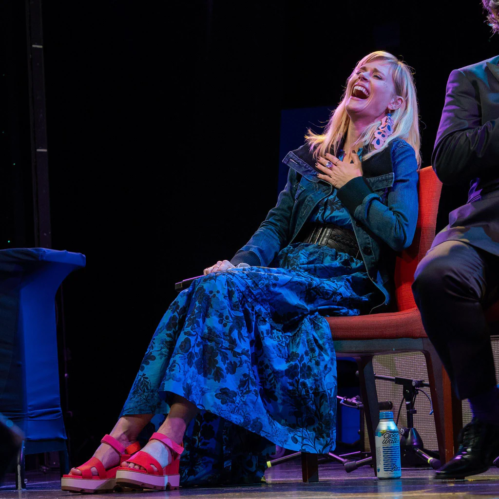 A woman laughing and holding her chest while seated on a stage during JoCo Cruise 2025, with a water bottle on the floor beside her.