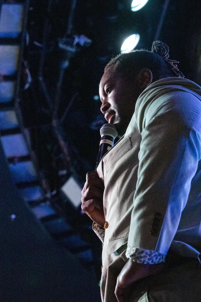 A person holding a microphone and speaking on stage during JoCo Cruise 2025.