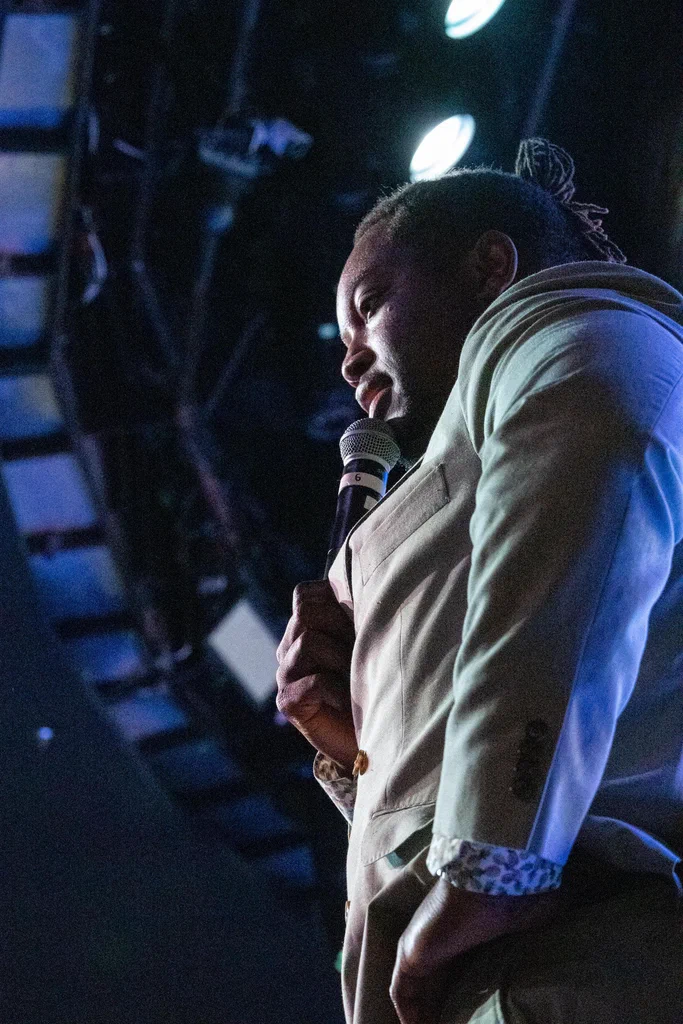 A person holding a microphone and speaking on stage during JoCo Cruise 2025.
