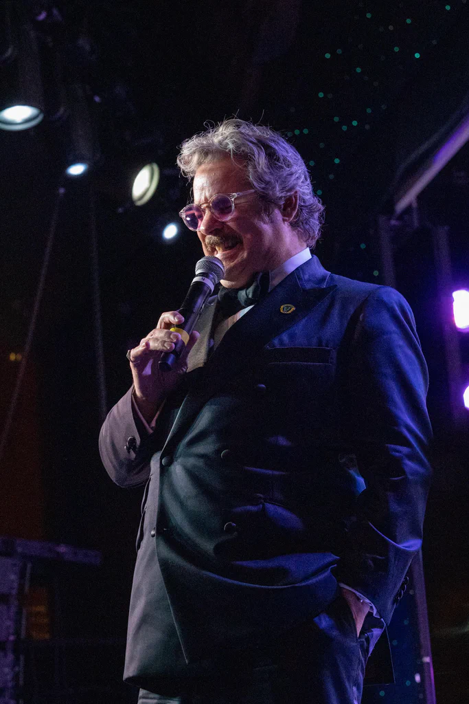 A man with curly gray hair and glasses, dressed in a suit, holds a microphone and appears to be performing or speaking on stage at JoCo Cruise 2025.