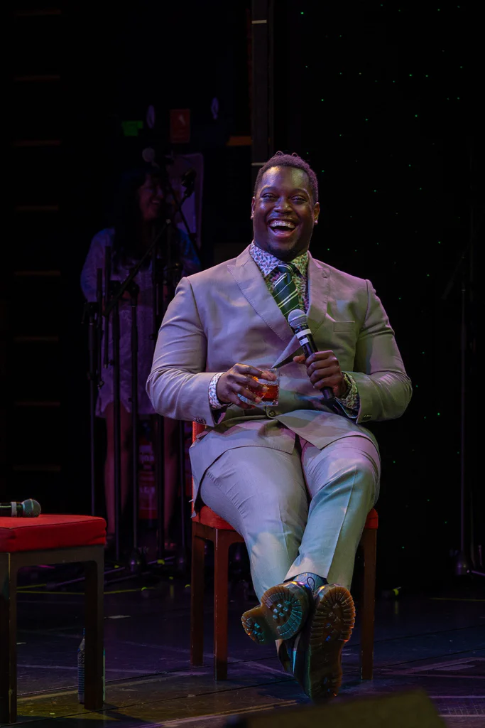 A man in a beige suit smiling and holding a microphone and a drink on stage during JoCo Cruise 2025.