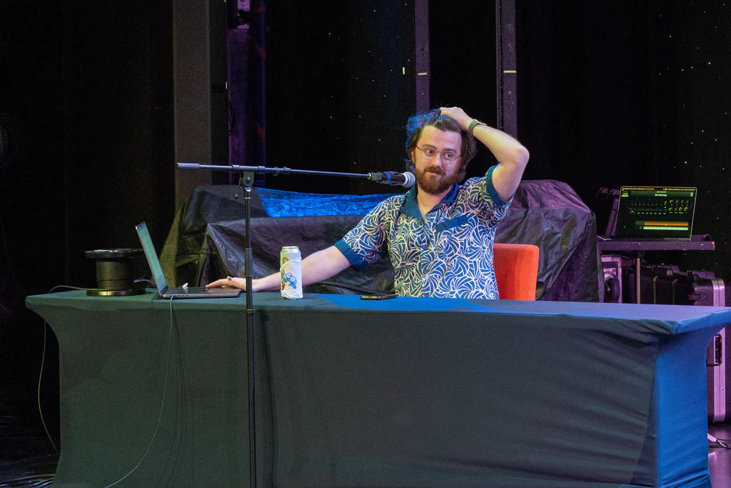 A person with glasses and a beard sitting at a table on a stage, with equipment and a microphone, touching their head while looking contemplative.