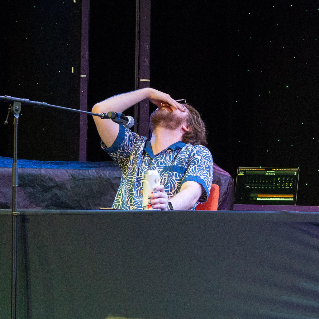 A person on stage at JoCo Cruise 2025, holding a drink, with their head tilted back and hand covering their face, possibly feeling overwhelmed or amused.