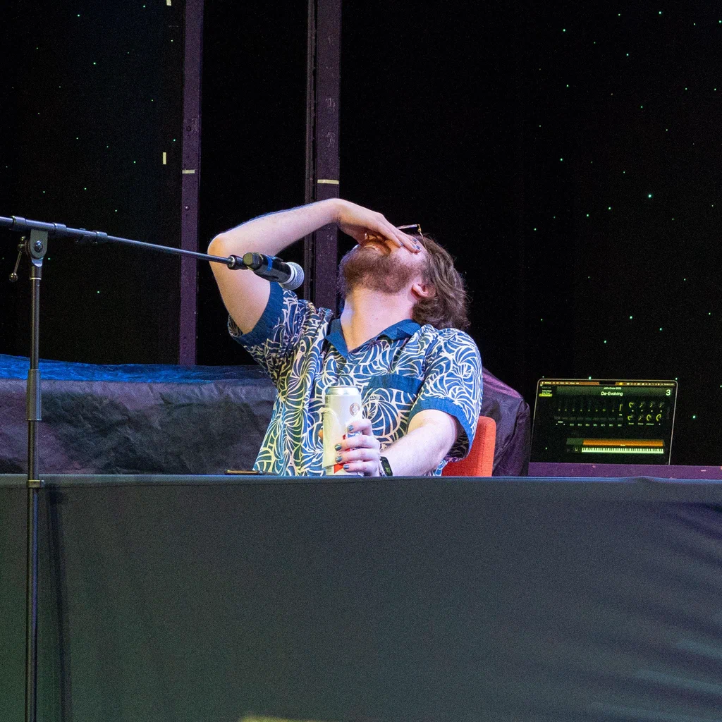 A person on stage at JoCo Cruise 2025, holding a drink, with their head tilted back and hand covering their face, possibly feeling overwhelmed or amused.