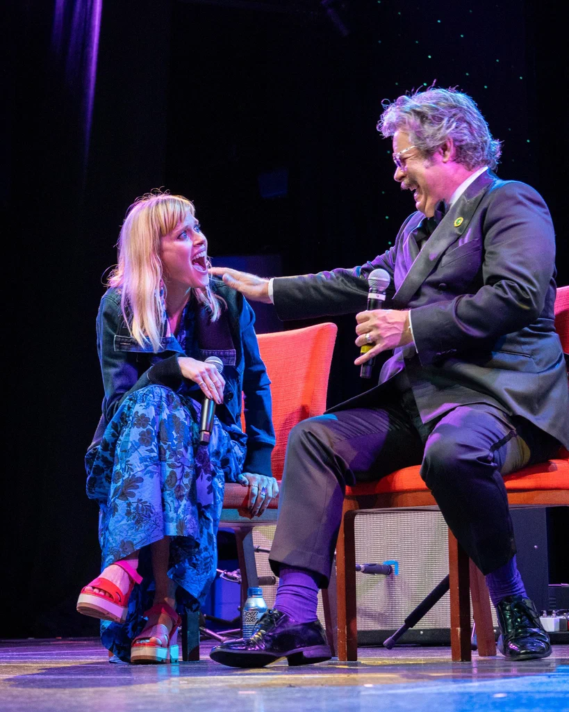 A woman and a man on stage during a comedy performance, engaging in a humorous interaction, both laughing. The woman is sitting on a chair holding a microphone, and the man is seated, touching her face with one hand, holding a microphone in the other.