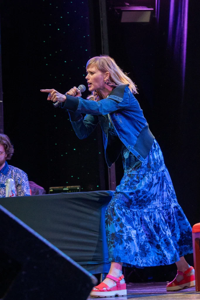 A woman on stage, holding a microphone, pointing and speaking passionately during a JoCo Cruise 2025 event.