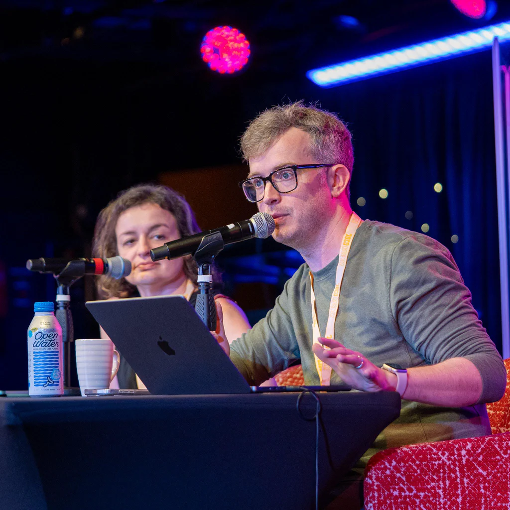 A man and a woman sit at a table with microphones, participating in a podcast during JoCo Cruise 2025, with a colorful stage background.