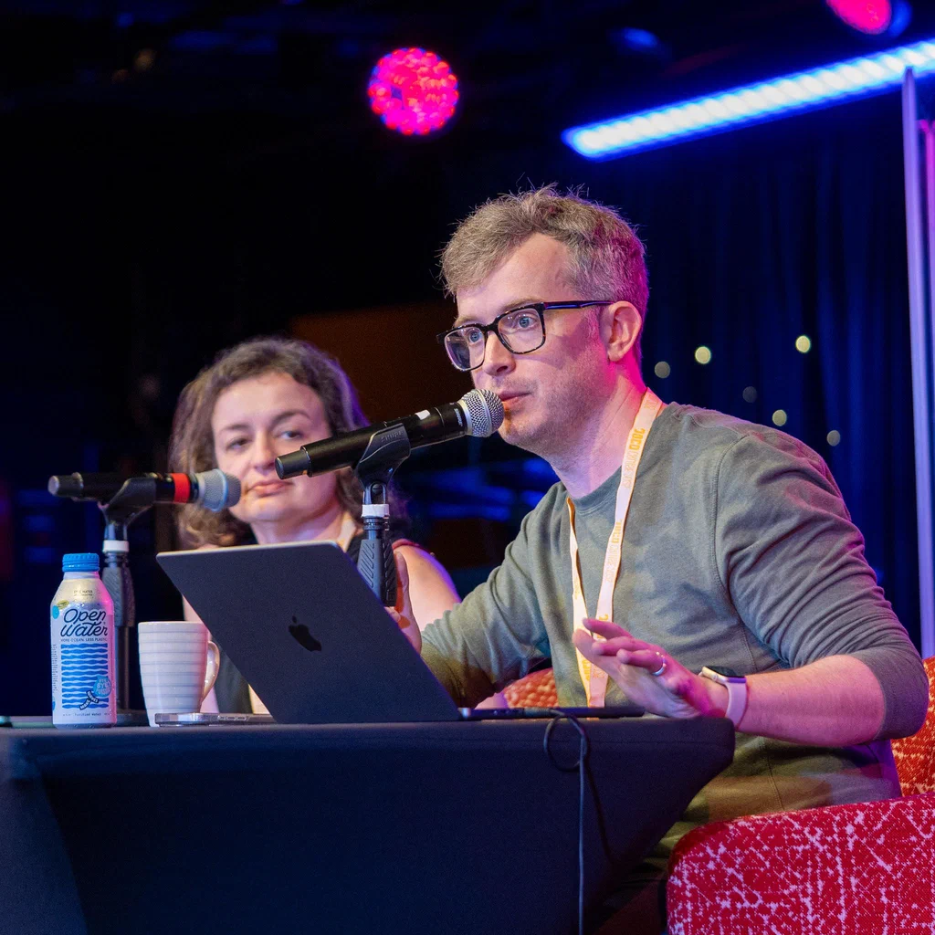 A man and a woman sit at a table with microphones, participating in a podcast during JoCo Cruise 2025, with a colorful stage background.