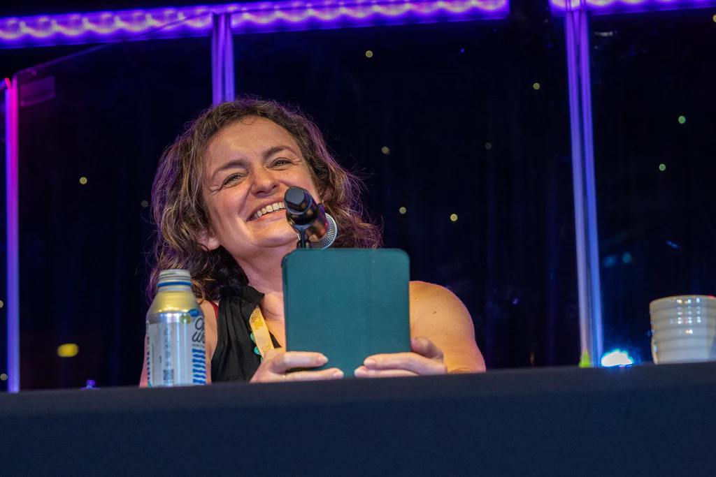 A woman smiling and speaking into a microphone during a podcast recording at JoCo Cruise 2025, with a can of soda and a stack of cups on the table in front of her.