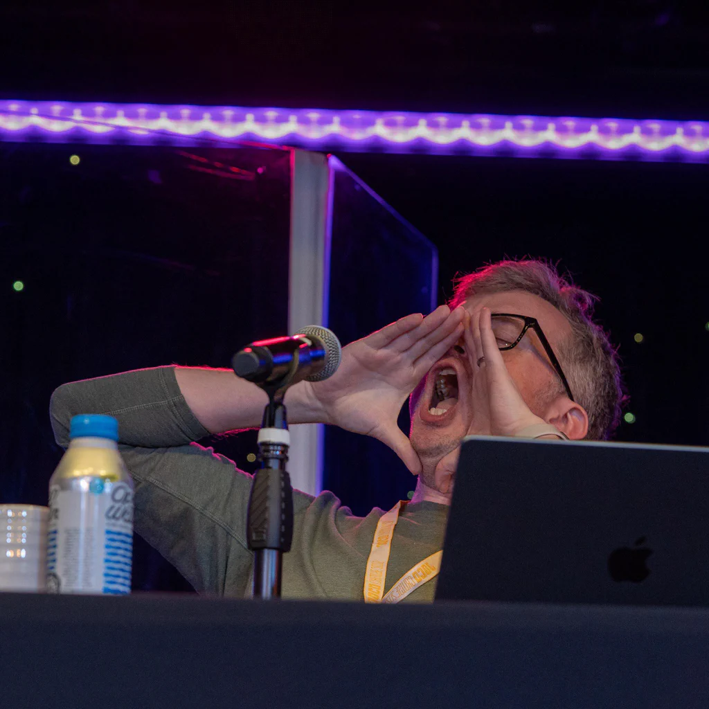 Person shouting or announcing at a microphone during JoCo Cruise 2025, with a laptop and drinks nearby.