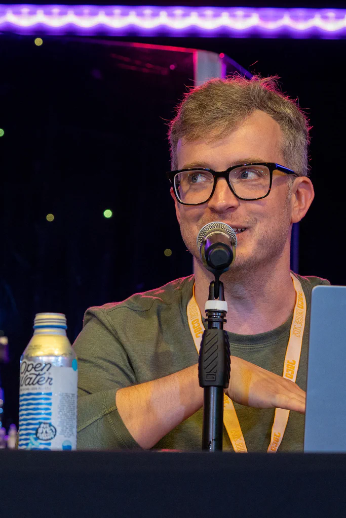 A man with glasses speaking into a microphone during a podcast on JoCo Cruise 2025.
