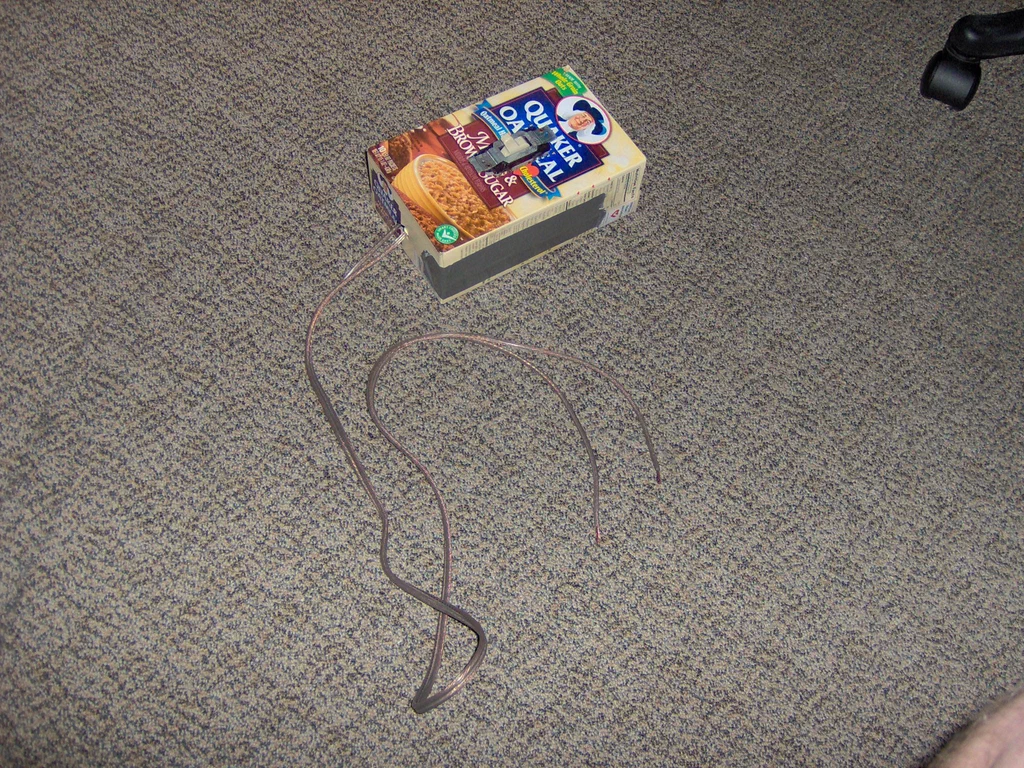 A high-voltage device sparking, with visible electrical arcs connecting metal wires on a carpeted floor near a cereal box.