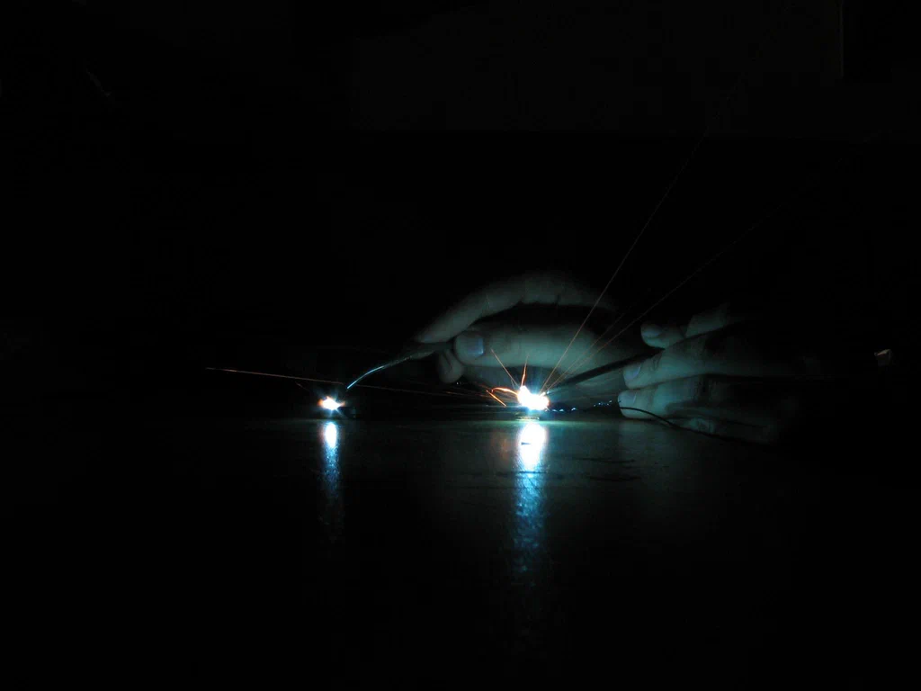 A person performing high-voltage welding or electrical work, with visible sparks and bright arcs of electricity against a dark background.