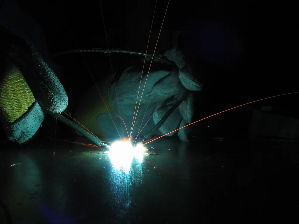 A person wearing protective gloves welds a high-voltage device, generating bright sparks and electrical arcs in a dark environment.