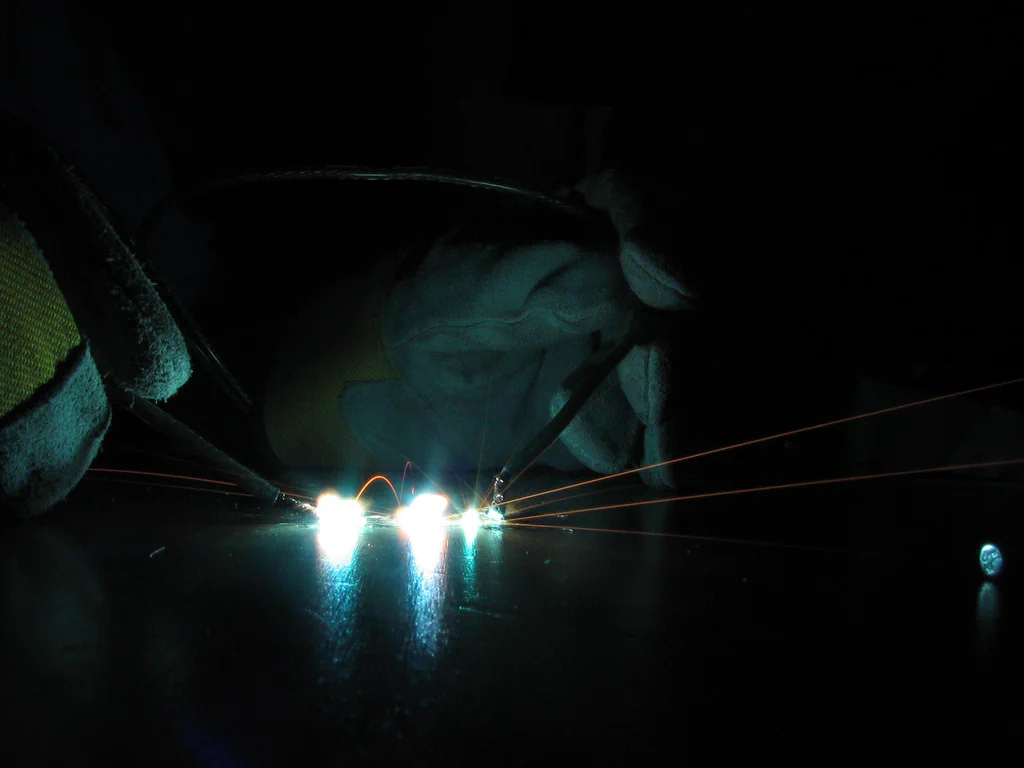 A person wearing gloves and protective eyewear is welding a high-voltage device, creating sparks and bright blue light in a dark environment.