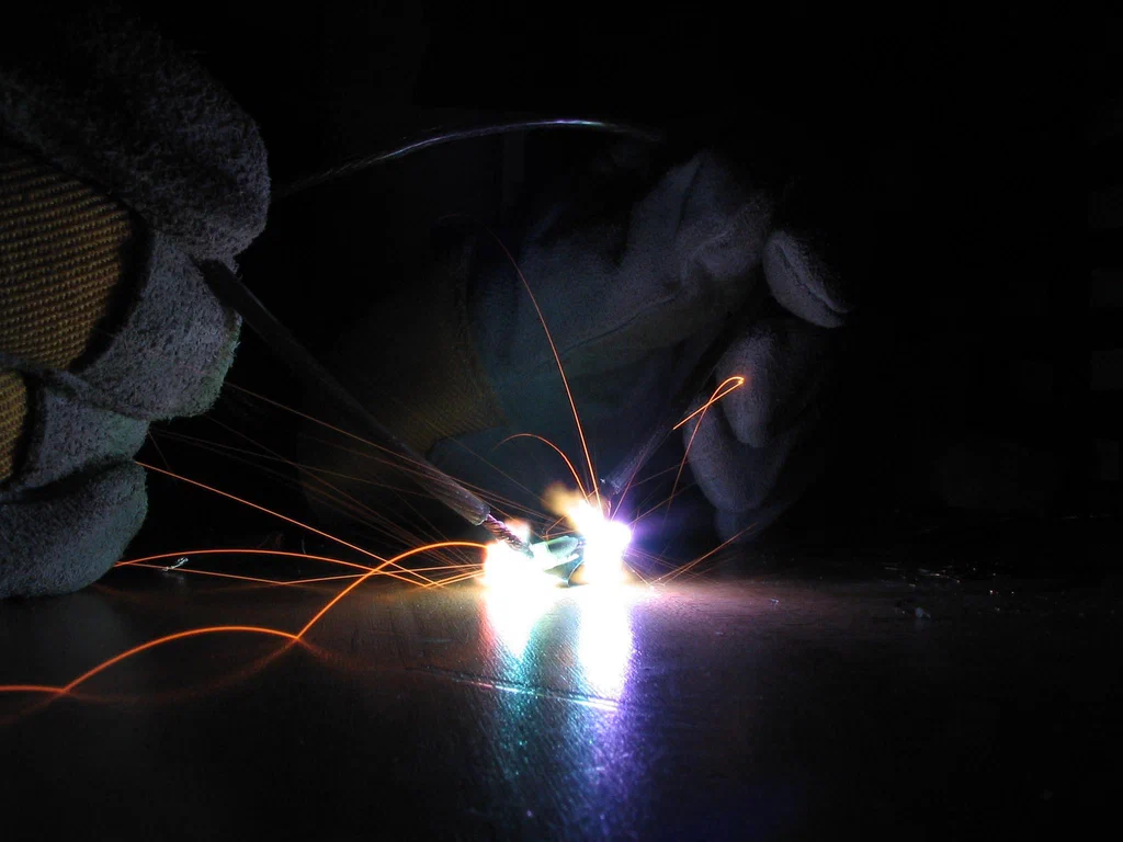 A close-up of a high-voltage device sparking, with sparks flying in various directions and a person wearing gloves working on the device in a dark environment.