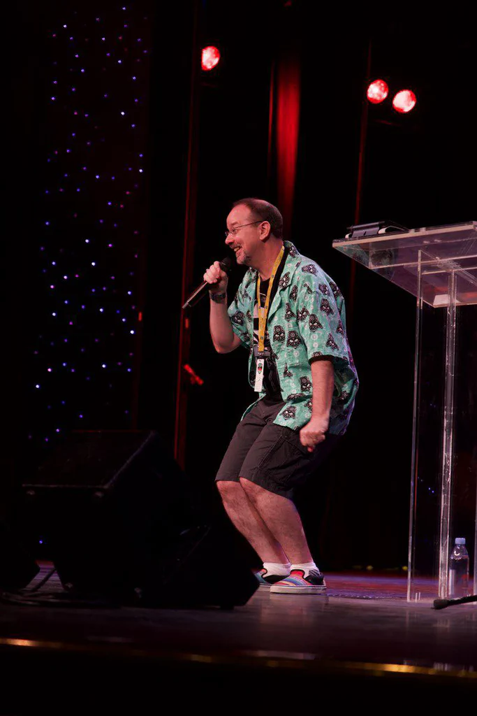 A man on stage holding a microphone, smiling and dancing, wearing a colorful Hawaiian shirt, shorts, and sneakers, with stage lighting and a transparent podium nearby.