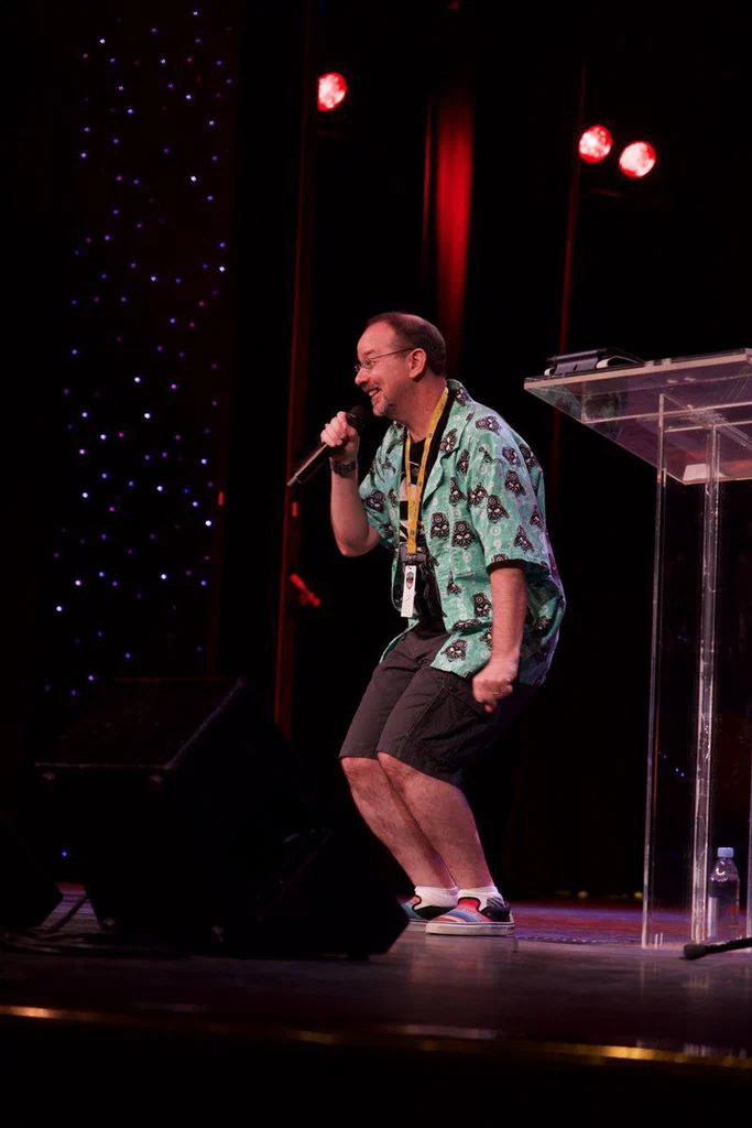 A man on stage holding a microphone, smiling and dancing, wearing a colorful Hawaiian shirt, shorts, and sneakers, with stage lighting and a transparent podium nearby.