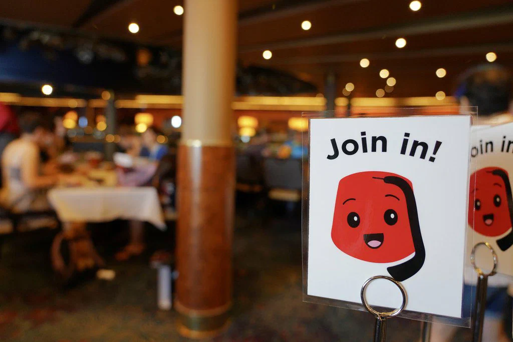 A sign with a cartoon red jelly character saying "Join in!" is placed inside a restaurant. The background shows diners sitting at tables with warm lighting and a busy atmosphere.