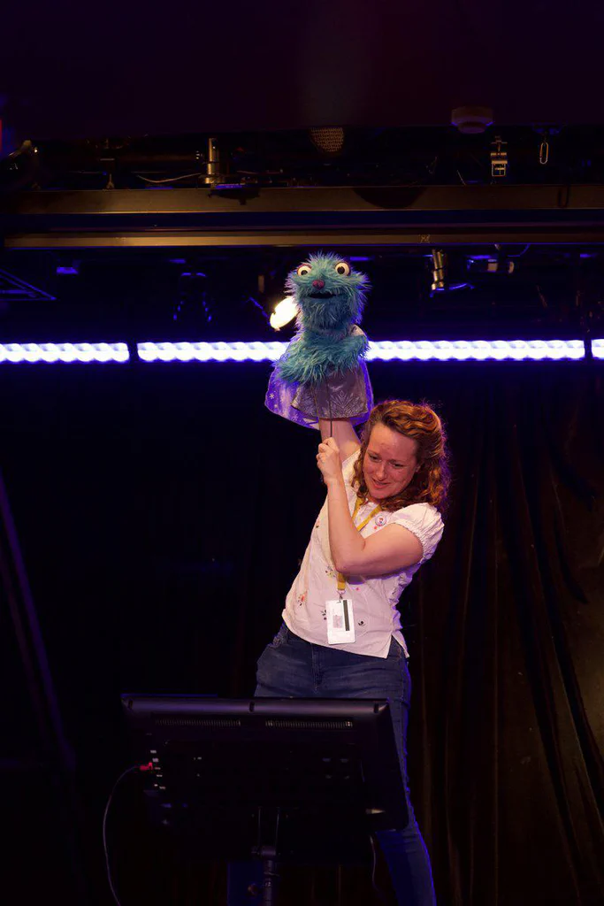 A woman holding a blue puppet with large eyes and a purple dress on a stage with black curtains and purple lighting.