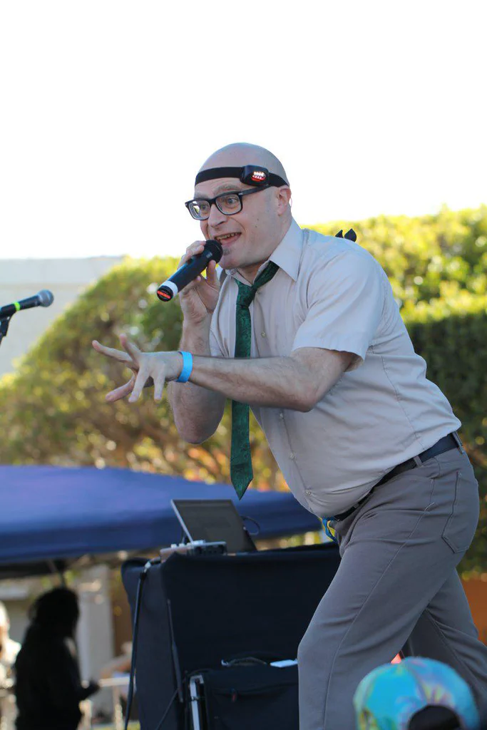 A man wearing glasses and a headlamp, holding a microphone, appears to be performing or speaking outdoors during daytime.