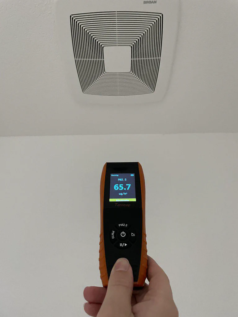 A hand holding a digital air quality monitor displaying a PM2.5 measurement of 65.7 µg/m³, with an exhaust vent on the ceiling above.