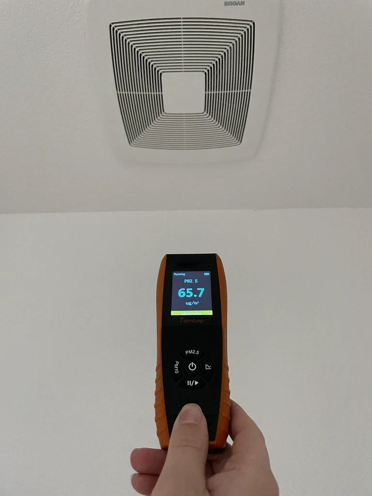 A hand holding a digital air quality monitor displaying a PM2.5 measurement of 65.7 µg/m³, with an exhaust vent on the ceiling above.