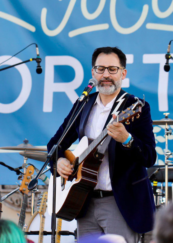 A man with glasses and a beard is playing an acoustic guitar and singing into a microphone during a live outdoor performance.