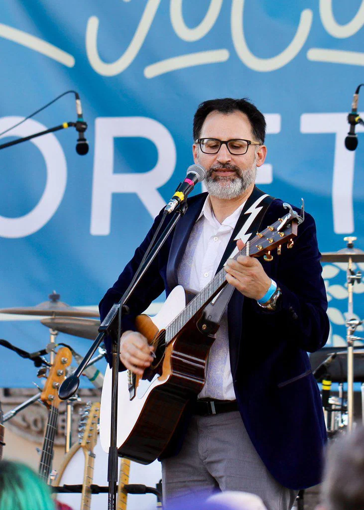 A man with glasses and a beard is playing an acoustic guitar and singing into a microphone during a live outdoor performance.