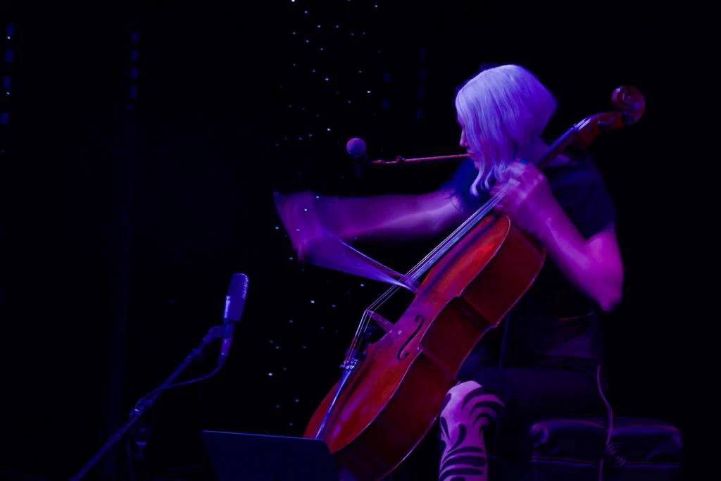 A woman with blonde hair playing a cello on a dark stage with purple lighting.