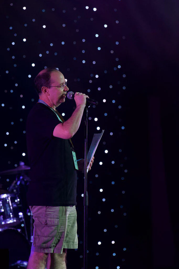 A man with glasses, holding a microphone and reading from a sheet of paper, standing on a stage with a starry background.