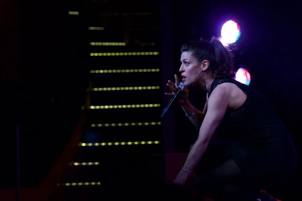 A woman crouching on stage, holding a microphone, illuminated by colorful stage lights.